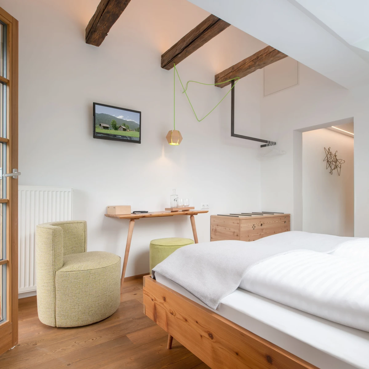 Schlafzimmer mit Holzbalken, Bett mit weißer Bettwäsche, Holzboden, Schreibtisch, grüne Drahtskulptur, Fernseher an der Wand