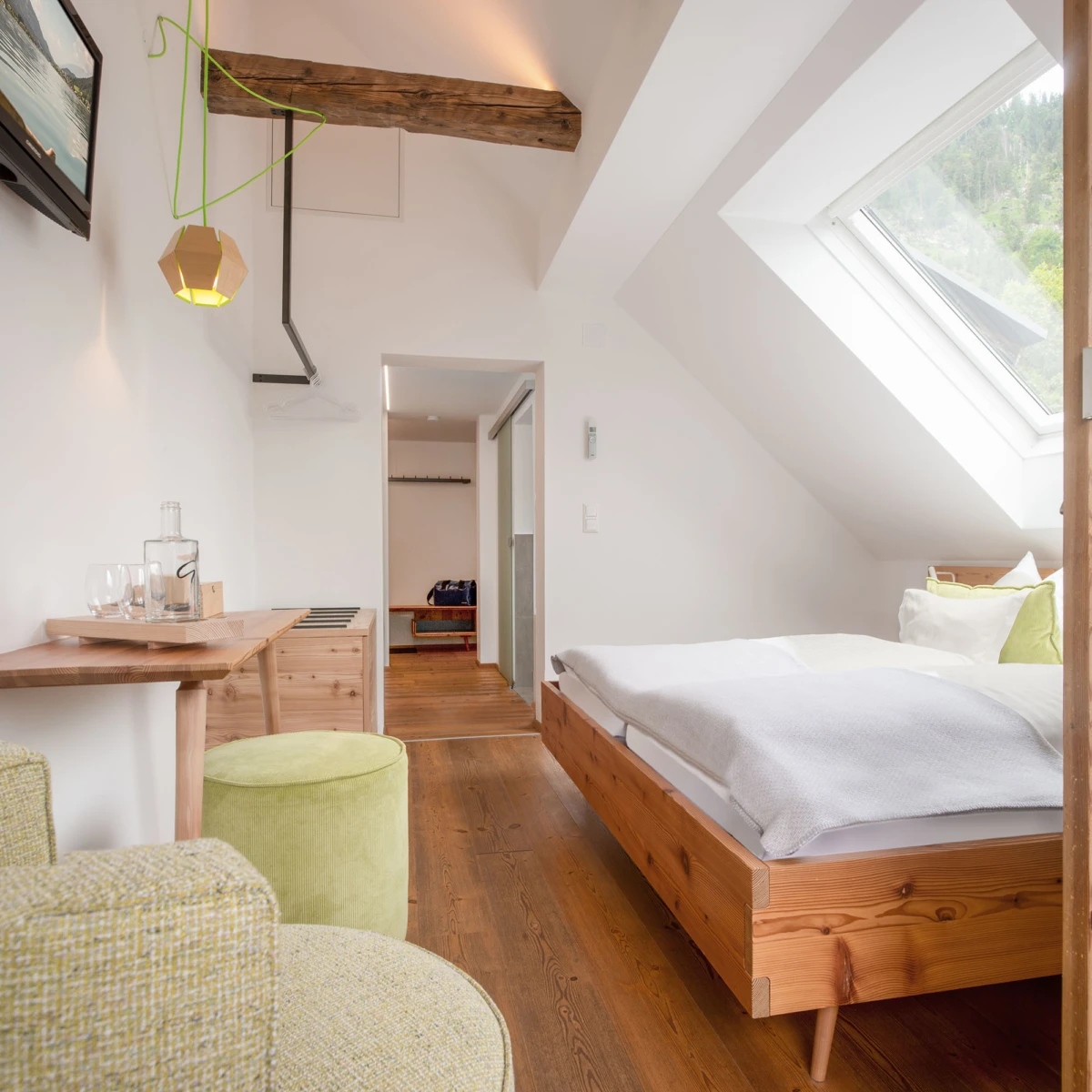 Dachgeschoss-Schlafzimmer mit massivem Holzbett, weißer Bettwäsche, Dachfenster, Holzfußboden, Arbeitsbereich und grünem Pouf.