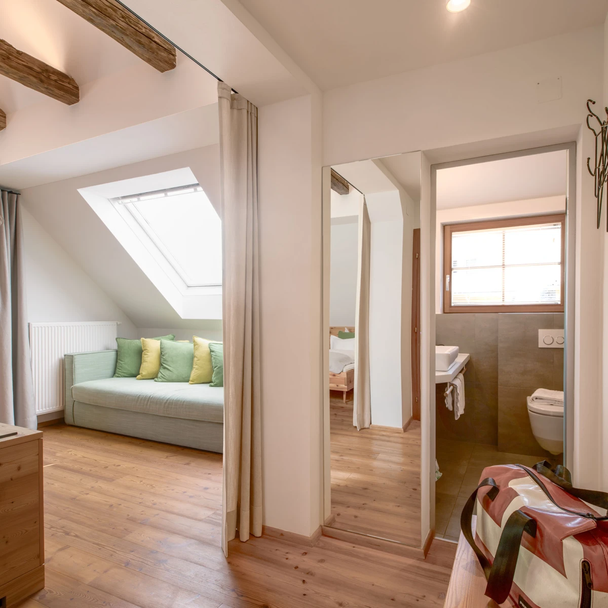 Dachgeschoss-Wohnraum mit grauem Sofa und gelben Kissen, Dachfenster, Vorhänge; Durchgang zum Badezimmer mit Waschbecken; Reisetasche im Vordergrund.