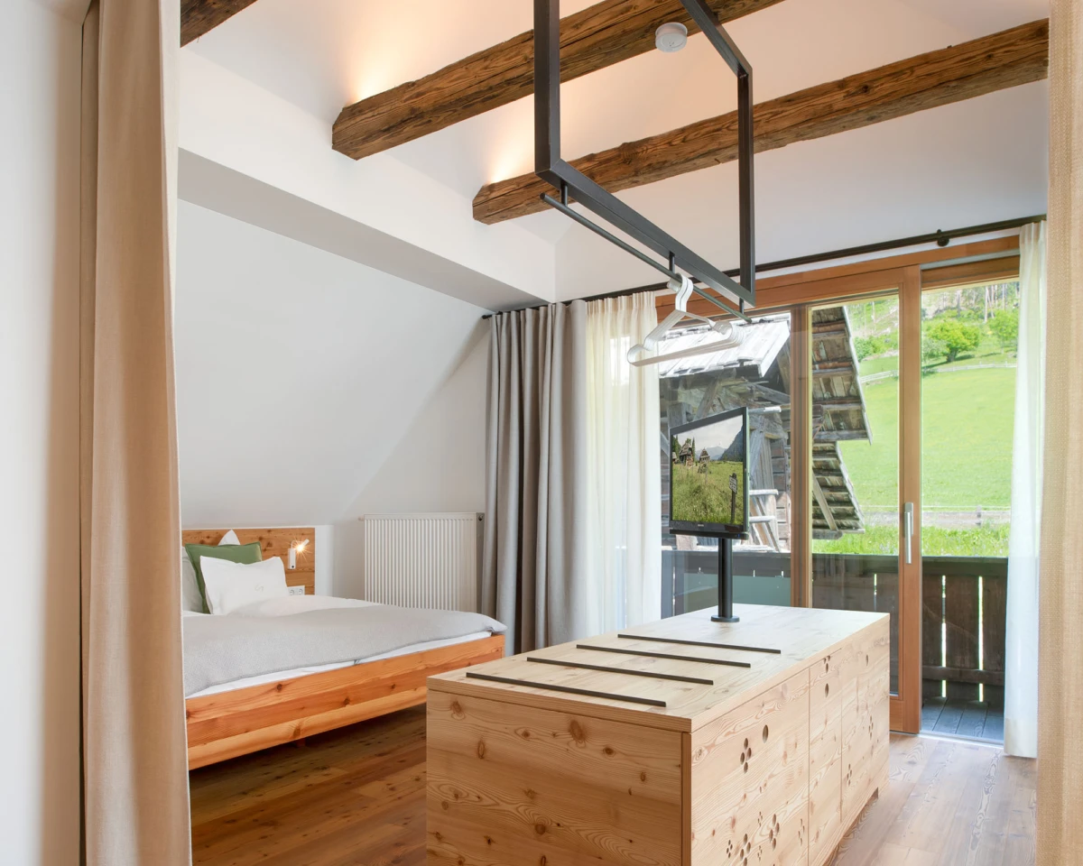 Schlafzimmer mit Holzbalkendecke, Holzbettenrahmen, graue Vorhänge, Heizkörper links, Holzkonsole mit Fernseher, Terrassentür zum grünen Garten
