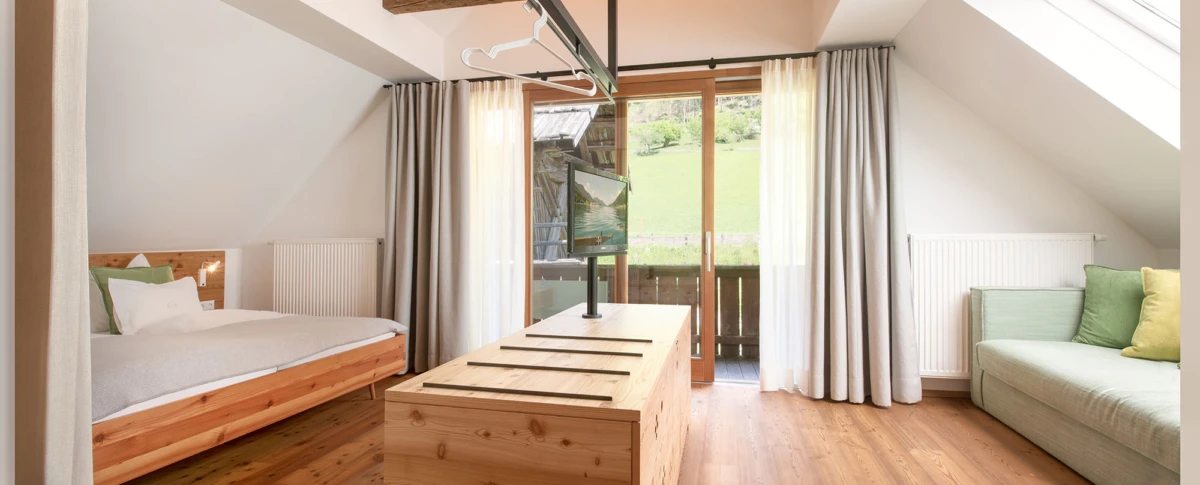 Helles Dachboden-Zimmer: Holzbett links, zentraler Holzkasten mit Fernseher, grüne Couch rechts, Glas-Türen zum Balkon mit Blick auf Wiese.