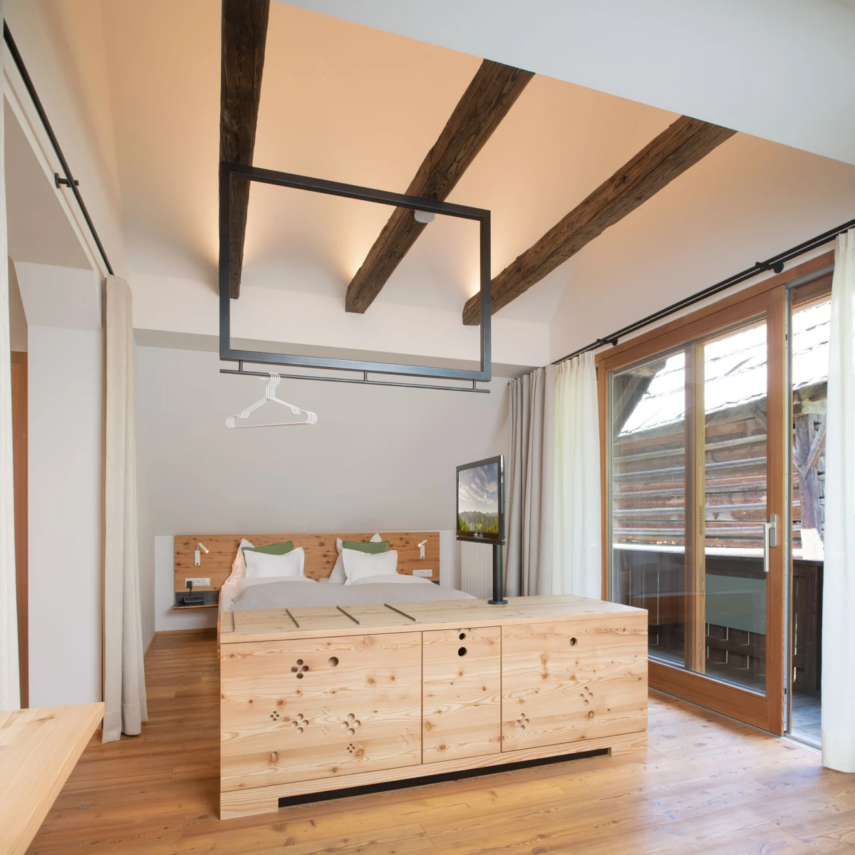 Schlafzimmer mit Holzbalkendecke, massiver Holzkasten als Raumteiler, Bett mit Bettwäsche in Weiß, TV und Glas-Schiebetür zum Balkon.