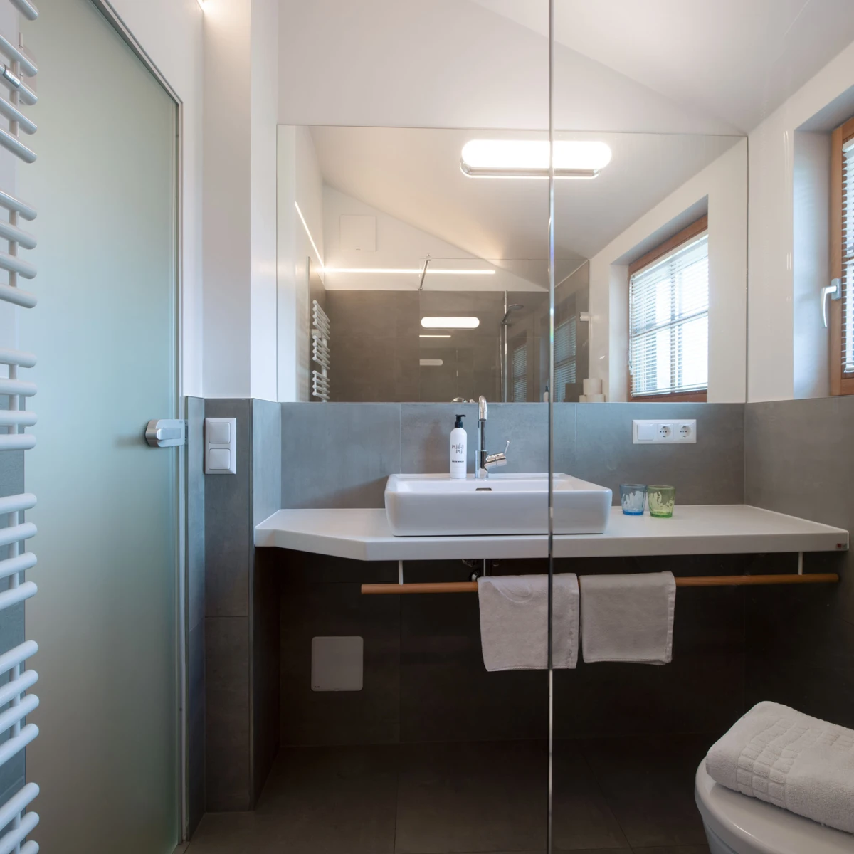 Waschbecken auf modernem Waschtisch im Badezimmer; Spiegel, Fenster, Handtücher, Dusche hinter Glas.