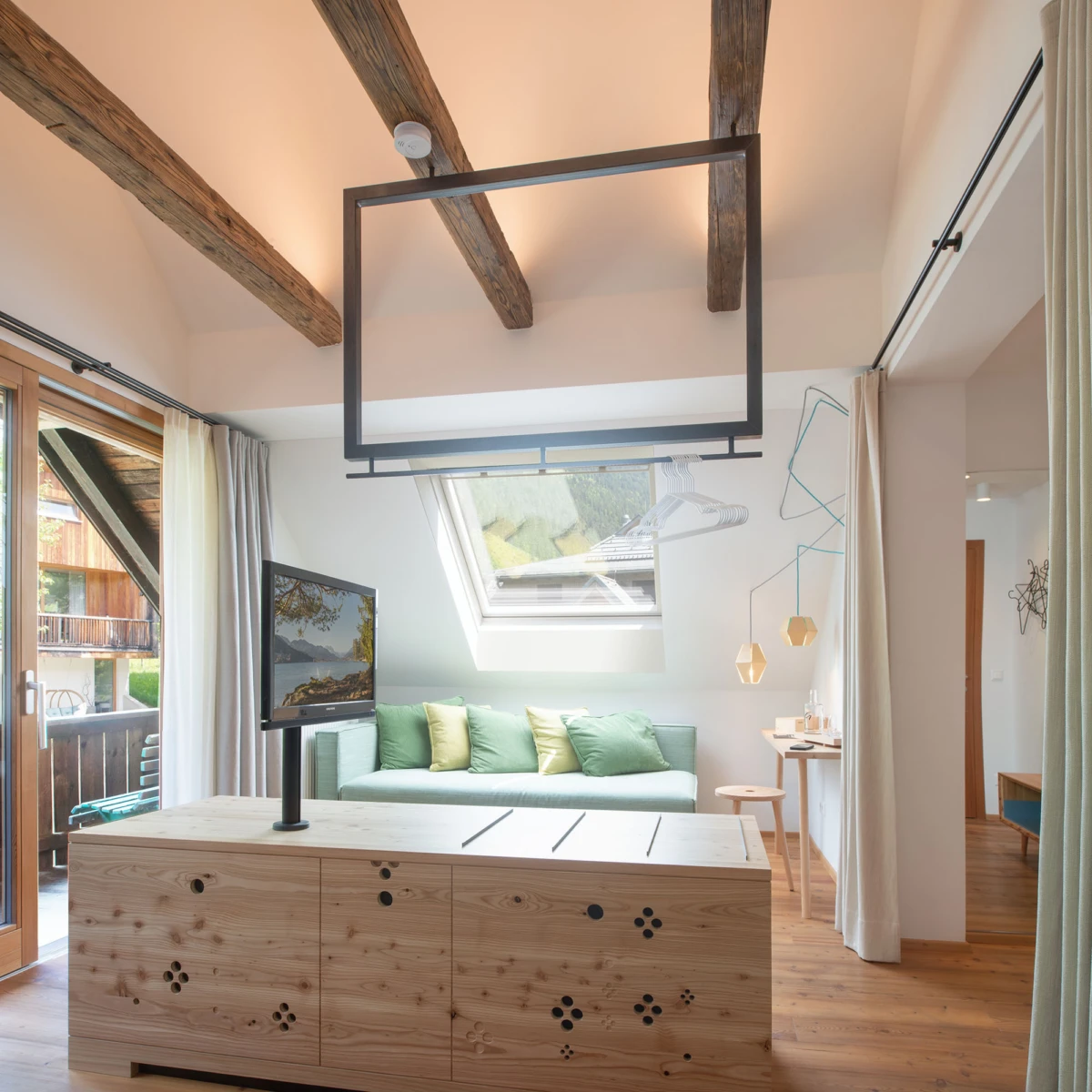 Helles Loft-Wohnzimmer mit Holzbalken, grünem Sofa, Monitor auf Holzkonsole; Dachfenster und Balkonblick