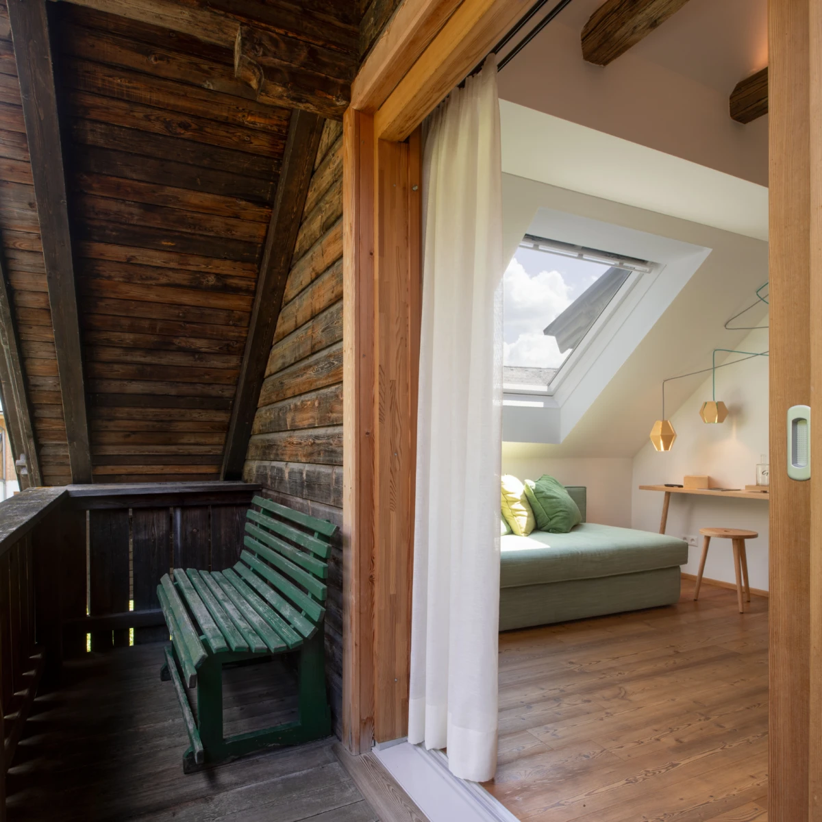 Grüne Holzbank auf dunklem Holzbalkon; Durchgang zum Schlafzimmer mit Bett, Schreibtisch und Dachfenster.